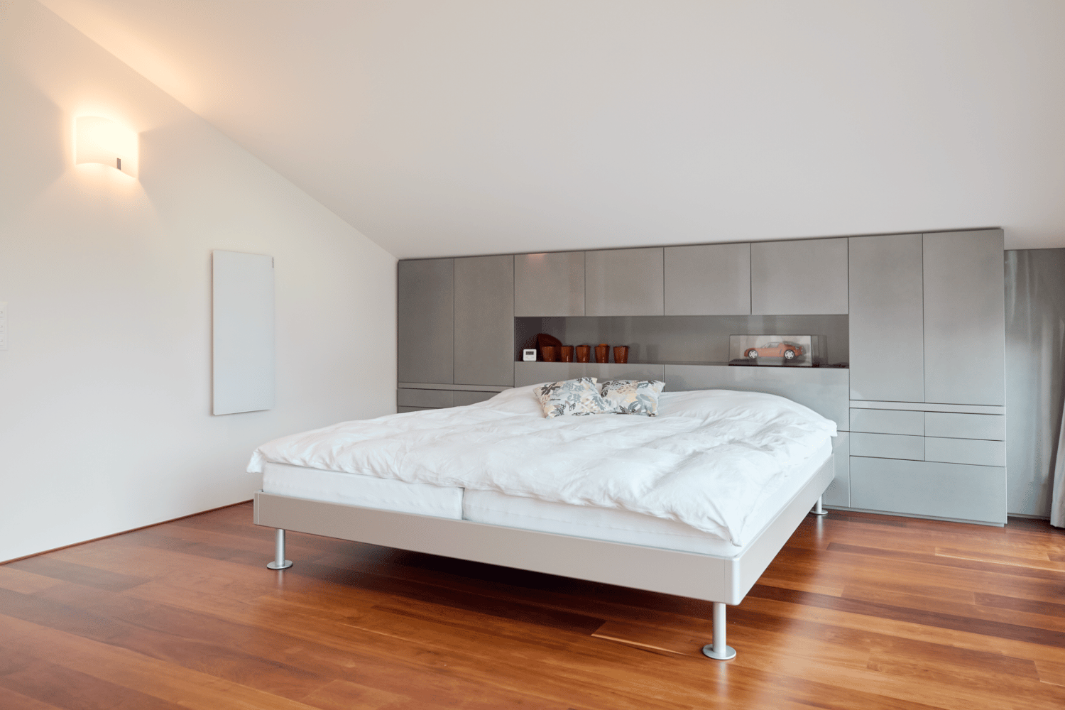 Eine weisse Infrarotheizung mit Glasoberfläche hängt an der Wand eines gemütlichen Schlafzimmers mit Bett und Nachttisch. 
