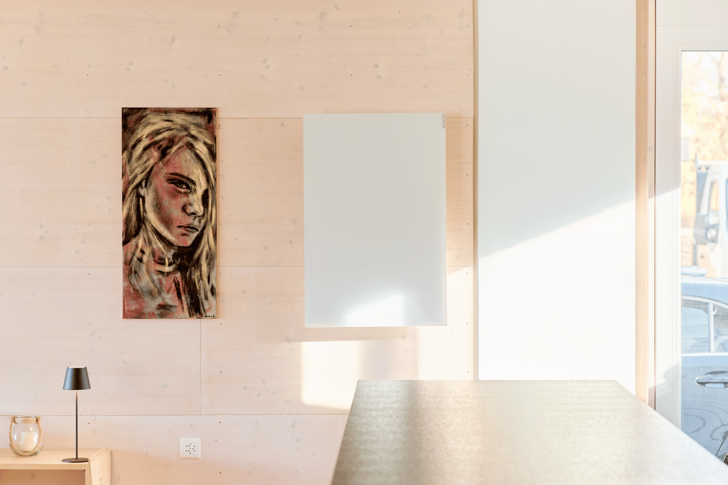 Eine weisse Infrarotheizung mit Glasoberfläche ist an der Wand eines modernen Wohnzimmers mit Sofa und einem grossen Fenster montiert.